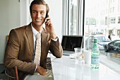 Junger Geschäftsmann mit braunem Sakko sitzt im Büro und telefoniert