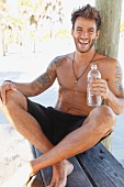 A young tattooed man sitting under a tree wearing shorts and holding a bottle of water