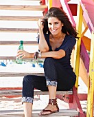 A young, dark haired woman sitting on a flight of wooden steps holding a bottle of water