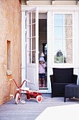 Vintage tricycle and outdoor furniture on wooden deck; child standing in open glass door