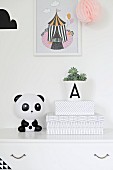 White box with graphic pattern and panda ornament on white chest of drawers below picture with circus motif on wall