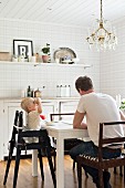 Father and child in high chair at white dining table below crystal chandelier