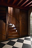 China crockery on steps of built-in spiral staircase in dark wood opening onto hallway with black and white chequered floor