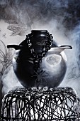 Düstere Inszenierung eines schwarzen Keramiktopfes, Kette mit Spinnenfigur und Hocker aus Metallgeflecht vor Rauchschwaden