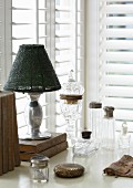 Antique glass vessels with silver lids, antiquarian books an table lamp in corner in front of windows with louvre blinds