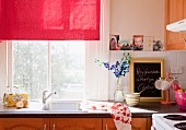 Window with red fabric roller blind above stainless steel sink in romantically decorated kitchen with wooden doors