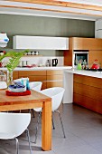 weiße Schalenstühle um Holztisch, im Hintergrund moderne Küchenzeile mit Massivholz Blenden an Unterschränken, grüngrau getönte Wand
