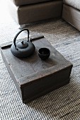 Holzkiste auf grau gesprenkeltem Webteppich mit schwarzer Teekanne und Becher