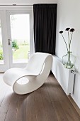 Modern, white Corian chaise in corner on wooden floor in front of dark curtains on French doors