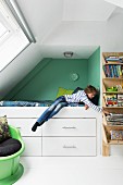 Teenager's bedroom with sloping ceiling; boy on raised bed in niche with drawers below