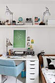 Blue, retro chair at white desk below table lamps and various objects on floating shelf in children's bedroom