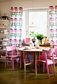 Pink wooden chairs around round table below window with floor-length, floral curtains