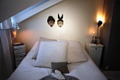 Bett vor Wand mit aufgehängten Masken, seitlich leuchtende Tischlampe auf Nachtkästchen in schlichtem Schlafzimmer im Dachgeschoss