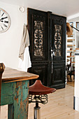 Bar stool with bicycle seat next to table and fitted cupboard with carved door panels made from dark wood
