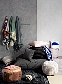 Verschiedene Sitzpoufs und Kissen auf Boden, vor grauer Wand mit Tüchern auf Hakenleiste