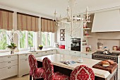 White country-house kitchen with free-standing counter and bar stools upholstered in red and white brocade below white chandelier