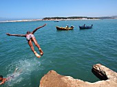 A boy diving from a rock into the sea with fishing boats in the background, Larache, Morocco
