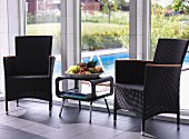 Rattan chairs flanking fruit bowl on retro side table in front of floor-to-ceiling windows with view of pool in garden