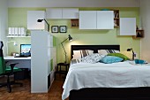 A room divider and wall cupboards hung at different heights to create lots of storage space between a double bed and a home office with a communal, green-painted wall