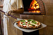 A pizza baker holding a margarita pizza on a paddle in front of a wood-fired oven
