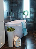 House-shaped lantern and floor vase in front of designer bathtub in dark-tiled bathroom