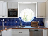 Porthole window above sink in Scandinavian kitchen with blue mosaic tiles