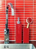 Seifenspender aus Keramik und Küchenbürste auf Edelstahlspüle mit moderner kantiger Armatur, rote Mosaikfliesen als Spritzschutz