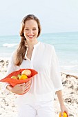 A young woman by the sea wearing a white blouse holding a hat filled with fruit