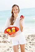 A young woman by the sea wearing a white blouse and shorts holding a hat filled with fruit