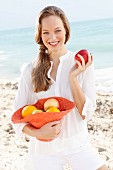 A young woman by the sea wearing a white blouse holding a hat of fruit