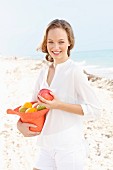 A young woman by the sea wearing a white blouse and shorts holding a hat filled with fruit