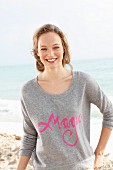 Junge, blonde Frau in grauem Pulli mit pinkfarbener Schrift am Meer