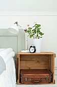Retro table lamp and geranium in white pot on wooden crate used as bedside table holding leather suitcase