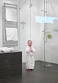 Kleines Kind im Bademantel vor der Glasabtrennung einer ebenengleichen Dusche, Handtuch-Heizkörper im Hintergrund