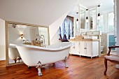 Freistehende Badewanne auf versilberten Klauenfüßen vor gerahmtem Spiegel auf Holzboden in grosszügigem Bad, im Hintergrund Doppelwaschtisch
