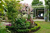 Round bed edged by low hedge in garden in front of garden swing in metal pergola