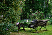 Parkbank, Tisch und Holzstühle auf Rasen in parkähnlichem Garten mit blühenden Stauden