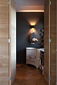 Open double door in wooden wall with view of cot against dark grey wall with sconce lamp