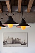 Lit, vintage pendant lamps below exposed, rustic roof beams, concrete girder and photograph on wall in loft apartment