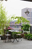 Alter Bistrotisch und schlichte Stühle vor Pflanzentöpfen auf Terrasse eines Lofts, Blick über Geländer auf die Stadt