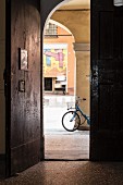 Hauseingang mit rustikaler Holztür und Blick in Arkadengang mit abgestelltem Fahrrad, gegenüber Geschäft mit bemaltem Rolladen