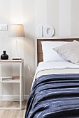 Table lamp on delicate, white bedside table next to bed against white and grey striped wallpaper