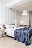 Schlafzimmer mit weißer Streifentapete, Doppelbett mit blauer Tagesdecke, Schreibplatz und verschiedenen Leuchten