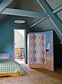 Painted screen next to bed and open door with view of chairs in adjoining room in converted attic with wood panelling painted blue-grey