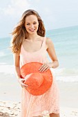 Fröhliche, junge Frau in apricotfarbenem Spitzenkleid und mit Hut in der Hand am Meer
