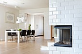 White dining table and black, upholstered chairs in modern, open-plan interior with corner fireplace in white-tiled wall