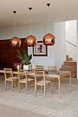 Long designer dining table and chairs made from pale wood under copper-coloured lampshades in modern dining area