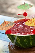 A tropical drink served in a halved watermelon with ice cubes and cocktail umbrellas