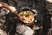 Jugendlicher bereitet Pfannengericht über Campingfeuer zu