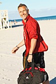 A middle-aged man on a beach wearing a red shirt and bathing shorts holding a sports bag